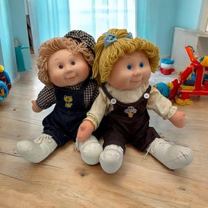 May include: Two vintage Cabbage Patch Kids dolls. One doll wears a black and white checked hat, denim dungarees, and white shoes. The other doll has blonde yarn hair, brown dungarees, and white shoes. Both dolls have freckles and blue eyes.
