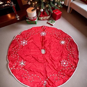May include: A bright red Christmas tree skirt with white snowflake embroidery and a white border. The skirt is circular with a hole in the center for the tree trunk. Festive holiday decor.