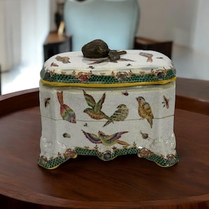 Könnte beinhalten: Dekorative Keramikbox mit Deckel, verziert mit handgemalten Vögeln und Insekten. Die Box ist weiß mit Craquelé-Finish, akzentuiert mit grünen und gelben Verzierungen. Ein bronzefarbener Knopf und Vogel schmücken den Deckel.