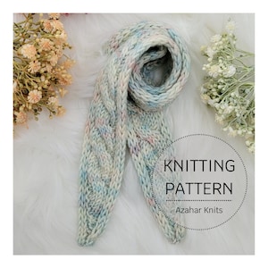 May include: A light blue and pink knitted scarf with a cable knit pattern. The scarf is folded in half and laid on a white fluffy surface. The text "KNITTING PATTERN" and "Azarhar Knits" is visible in the image.