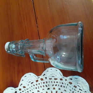 Op de afbeelding: Een heldere glazen fles met een metalen schroefdop en een handvat. De fles heeft een lichtblauwe tint en staat op een houten oppervlak.