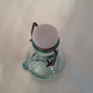 Op de afbeelding: Een vintage glazen fles met een witte keramische dop en een metalen draad sluiting. De fles is helder en heeft een groene tint.