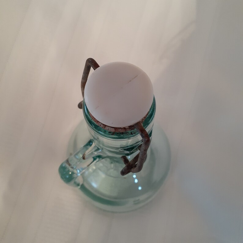Op de afbeelding: Een vintage glazen fles met een witte keramische dop en een metalen draad sluiting. De fles is helder en heeft een groene tint.