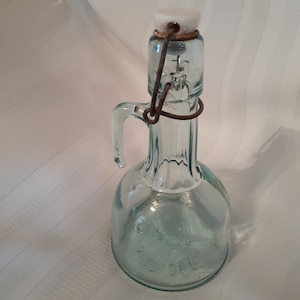 Op de afbeelding: Een vintage glazen fles met een draadbeugel sluiting en een witte keramische stop. De fles is helder en heeft een getextureerd oppervlak.