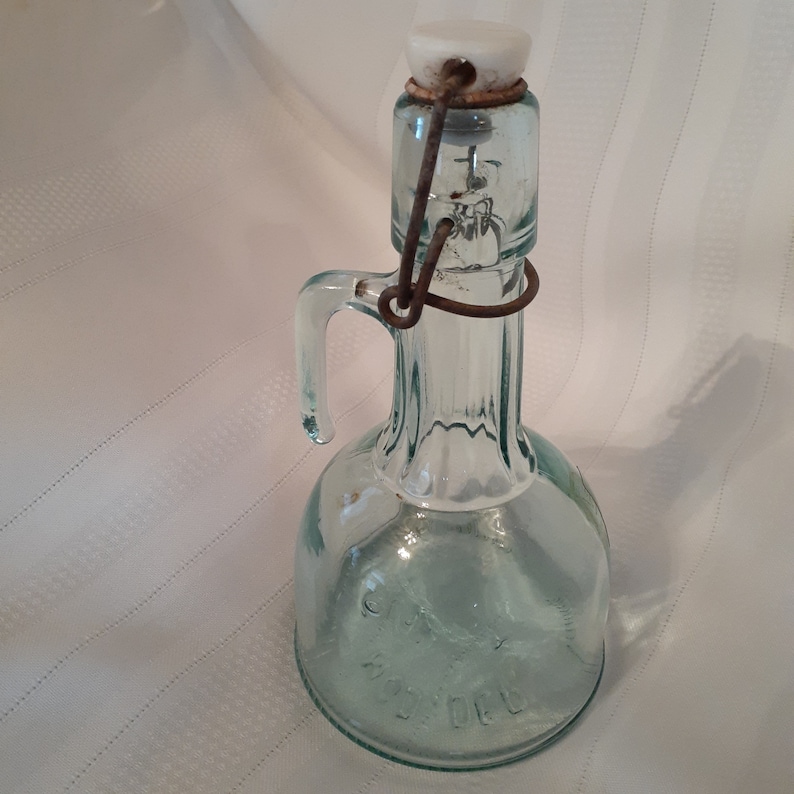 Op de afbeelding: Een vintage glazen fles met een draadbeugel sluiting en een witte keramische stop. De fles is helder en heeft een getextureerd oppervlak.