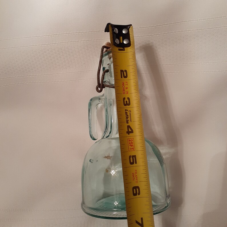 Op de afbeelding: Een heldere glazen fles met een metalen handvat en een kurken stop. De fles is ongeveer 15 cm hoog en heeft een ronde bodem. Hij staat op een wit oppervlak.