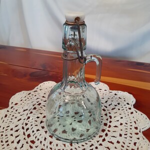 Op de afbeelding: Een vintage glazen fles met een scharnierend metalen deksel en een witte porseleinen stop. De fles heeft een heldere, groene tint en een rond lichaam met een handvat.