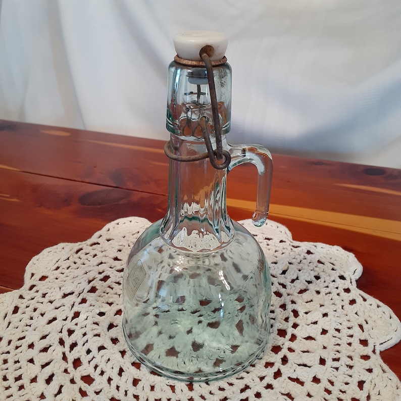 Op de afbeelding: Een vintage glazen fles met een scharnierend metalen deksel en een witte porseleinen stop. De fles heeft een heldere, groene tint en een rond lichaam met een handvat.