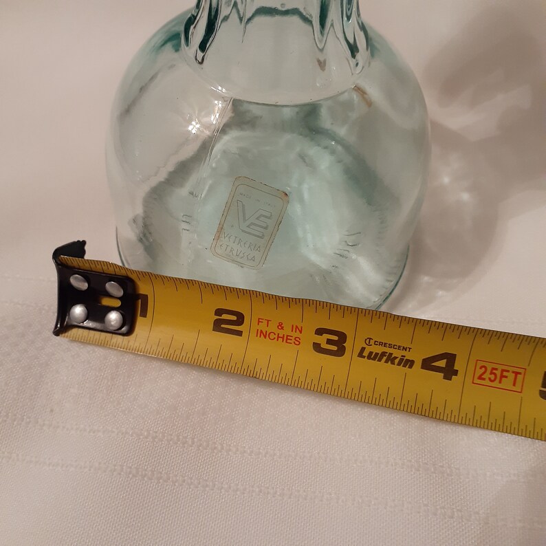 Op de afbeelding: Een doorzichtige glazen fles met een getextureerd oppervlak. De fles is ongeveer 7,6 cm hoog. De fles heeft een etiket met de tekst "Vitreka Vitreka".