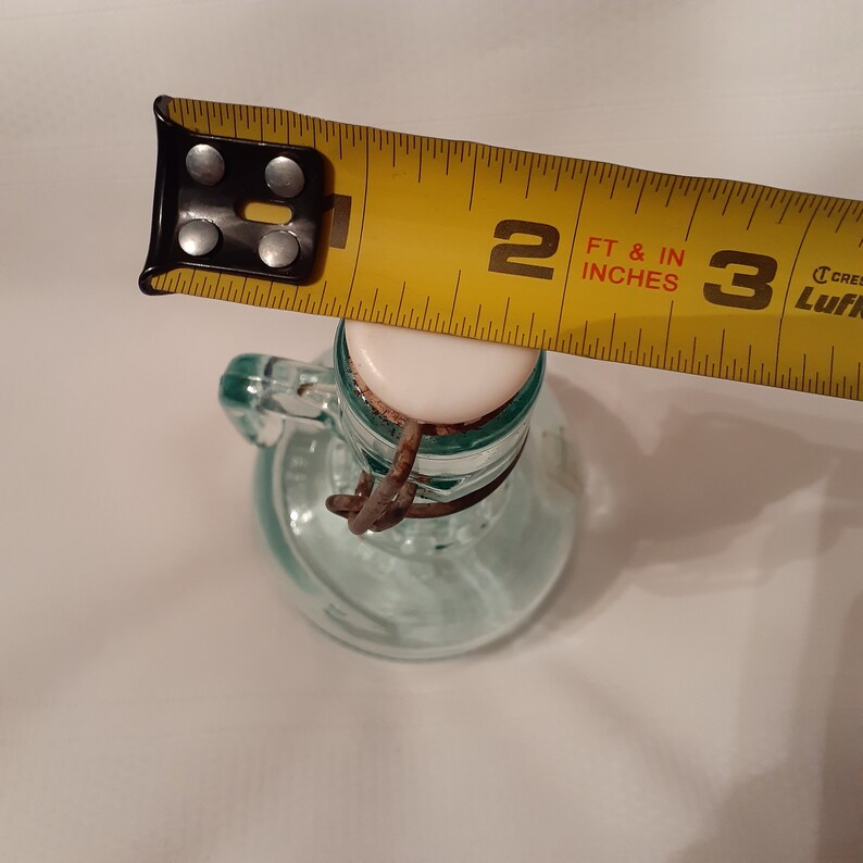 Op de afbeelding: Een doorzichtige glazen fles met een wit deksel en een roestige metalen sluiting. De fles is 5 cm hoog en wordt gemeten met een gele meetlint.