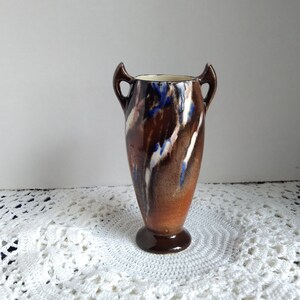 May include: A brown ceramic vase with blue and white abstract designs and two handles. The vase is sitting on a white crocheted doily.