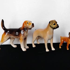 May include: Three ceramic dog ornaments. A brown and tan Beagle stands to the left, a tan Boxer in the middle, and a small orange Boxer on the right. All dogs are posed in a standing position.