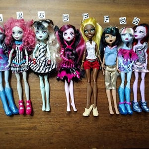 Peut inclure: Huit poupées Monster High avec des coiffures et des tenues différentes. Les poupées sont debout sur une surface en bois. Les poupées portent une variété de couleurs, notamment le rose, le bleu, le noir, le blanc et le rouge.