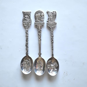 May include: Three silver Christmas spoons. Each spoon has a different embossed design, including angels and religious figures. The spoons are dated 1977, 1978, and 1979. The spoons have ornate handles and detailed bowls.
