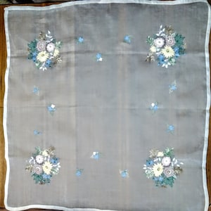 Peut inclure: Une nappe en tissu blanc transparent avec des motifs floraux brodés dans des tons de bleu, jaune et vert.