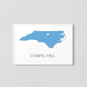 May include: A white framed print with a blue outline of the state of North Carolina. A small white heart is placed over Chapel Hill, North Carolina. The text "CHAPEL HILL" is printed below the state outline.