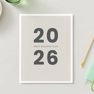 Op de afbeelding: Een ingelijste print met de tekst "2026 BIBLE READING PLAN" in donkergrijs. Een potlood, een klein groen notitieboekje en een sleutelbos staan rechts. Een kom met groene druiven en een glas vloeistof staan linksboven.
