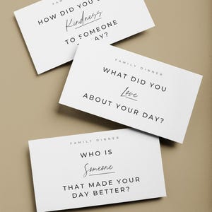 May include: Three white cards with black text for a family dinner game. The cards ask questions like "How did you show kindness today?" and "What did you love about your day?" on a neutral background.