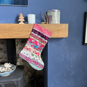 May include: A patchwork Christmas stocking with floral patterns in pink, red, and blue hangs from a wooden mantelpiece. The stocking is a festive decoration. A wooden Christmas tree and a white candle are on the mantelpiece, above a fireplace.