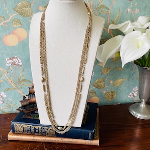 Op de afbeelding: Een vintage goudkleurige ketting met meerdere strengen fijne kettingen en decoratieve rechthoekige en ovale accenten. De ketting wordt getoond op een witte mannequin buste tegen een bloemen behang achtergrond.