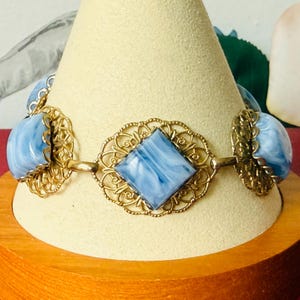 Puede incluir: Una pulsera con cabujones veteados en azul y blanco alternados, engastados en monturas de metal dorado con filigrana. La pieza central es una piedra veteada en forma de rombo rodeada por un marco de metal dorado calado y ornamentado.