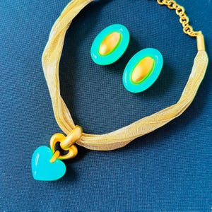 May include: A jewelry set consisting of a gold-toned ribbon choker necklace with a heart-shaped turquoise pendant and a pair of matching oval-shaped turquoise earrings with gold-toned centers, featuring a gold-toned chain extender.