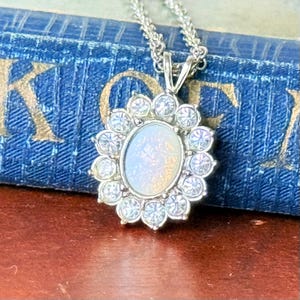 May include: A silver necklace with a pendant featuring an oval, iridescent stone surrounded by a halo of clear, round gemstones. The pendant is suspended from a delicate silver chain. The background is a blurred view of a blue book.