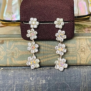 May include: A pair of long, dangling earrings with a silver chain and five clear, iridescent flower-shaped rhinestones.