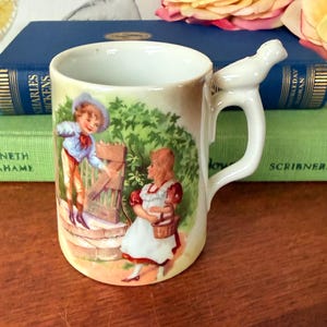 Puede incluir: Taza de porcelana vintage con asa en forma de pájaro. La taza presenta una colorida ilustración de dos niños cerca de una puerta de madera. El fondo es amarillo claro con follaje verde. La taza está sobre una superficie de madera.