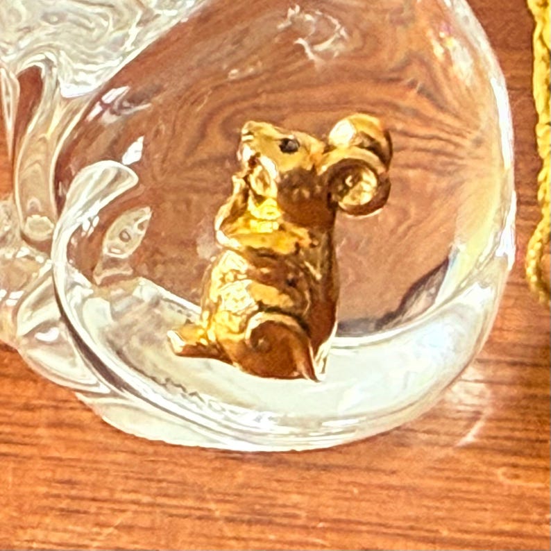 Puede incluir: Una figura de carnero dorada dentro de un cuenco de vidrio transparente. El carnero tiene cuernos rizados y una superficie texturizada. El cuenco tiene forma redondeada y est&aacute; sobre una superficie de madera.