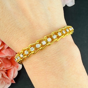 Puede incluir: Una pulsera de metal en tono dorado con un diseño de eslabones geométricos repetitivos. Cada eslabón está engastado con un único estrás transparente de corte redondo. La pulsera tiene un acabado pulido y una estructura flexible.