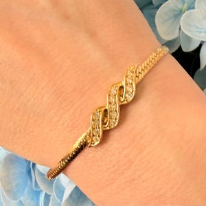 May include: A gold-toned bracelet with a textured, snake-like design. The bracelet features a central, intertwined pattern adorned with small, clear gemstones. The bracelet is worn on a wrist, with a soft, neutral skin tone.