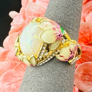 May include: A gold-tone ring featuring a large, iridescent oval opal-style cabochon center stone. The band is decorated with cream-colored enamel flower petals with pink accents and small, clear pavé-set crystals along the metal frame.