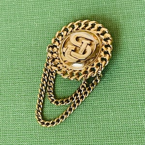 Könnte beinhalten: Eine goldfarbene Brosche mit einem runden Design, das die Buchstaben "GS" in einem erhabenen, ineinandergreifenden Muster zeigt. Die Brosche ist an einer doppelten Kette befestigt.