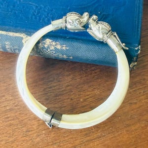 May include: Ivory-colored bangle bracelet with silver-toned elephant head accents. The bracelet features a hinged clasp and is resting on a wooden surface. The elephant heads are facing each other.