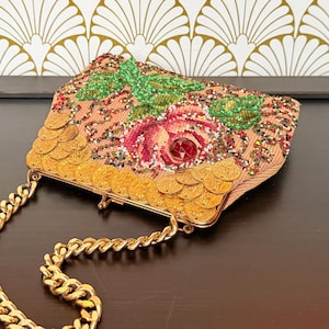 May include: A vintage-style handbag with a gold chain strap. The bag features a pink fabric base adorned with a floral design, green beaded leaves, and a large red gem. Gold coin-shaped embellishments line the frame.