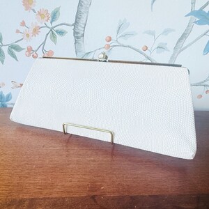 May include: A white rectangular clutch bag with a textured surface and a metallic gold-toned frame. The bag features a central clasp closure at the top, designed with a small, rounded decorative element. The overall shape is elongated and structured.
