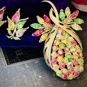 Pode incluir: Broche e brincos a condizer em estilo vintage, em tom dourado. O broche apresenta um design grande em forma de gota preenchido com strass iridescentes coloridos. Os brincos e o broche têm detalhes em rosa, verde e amarelo.