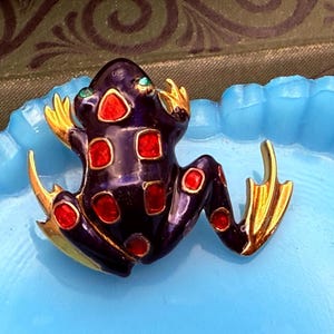 Puede incluir: Un broche de rana púrpura con detalles rojos y dorados. La rana tiene ojos verdes y está decorada con puntos y cuadrados rojos.