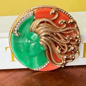 May include: A vintage brooch featuring a Medusa design. The brooch has a gold-tone frame with a green face on one side and a red background on the other. Gold snakes and clear rhinestones add detail.
