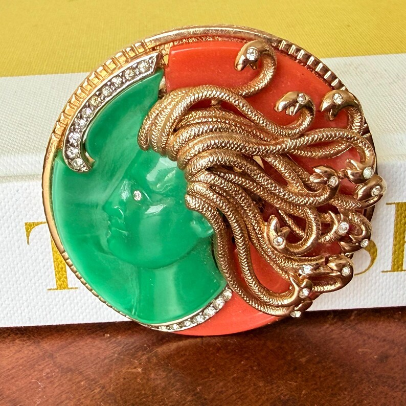 May include: A vintage brooch featuring a Medusa design. The brooch has a gold-tone frame with a green face on one side and a red background on the other. Gold snakes and clear rhinestones add detail.