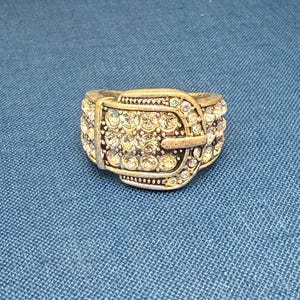May include: A silver-toned ring in the shape of a belt buckle, encrusted with numerous clear, sparkling rhinestones. The ring has a detailed design with a buckle and a textured band, set against a blue background.
