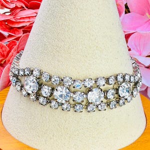 May include: A silver-tone metal bracelet featuring three rows of clear, round-cut rhinestones. The design alternates between smaller stones and larger, prominent circular stones set in a repeating pattern across the length of the bracelet.