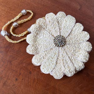 Può includere: Pochette a forma di margherita color crema, con manico di perline. I petali sono ornati da ricami e paillettes dettagliati. Il centro del fiore presenta strass iridescenti. Un accessorio di ispirazione vintage.