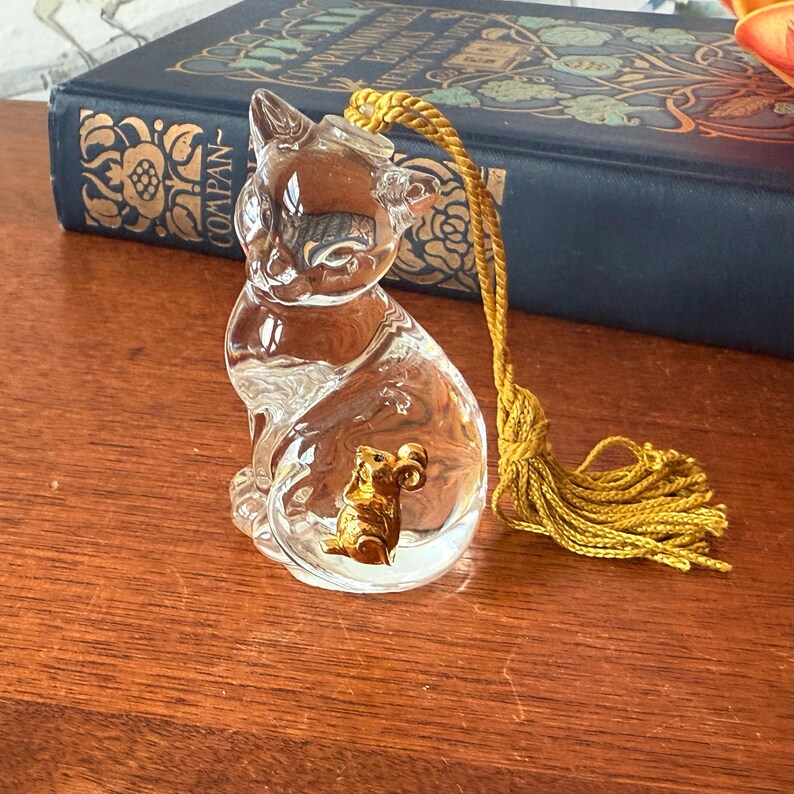Puede incluir: Una figura de gato de cristal transparente con un rat&oacute;n dorado en su interior. El gato tiene una borla amarilla atada a la cabeza. La figura est&aacute; sobre una superficie de madera junto a un libro azul.