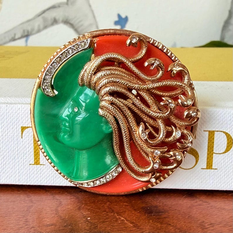 May include: A round brooch featuring a Medusa design. The brooch is divided into two sections: a green face with a detailed profile and an orange background with gold snakes. The brooch is trimmed with gold and small clear crystals.