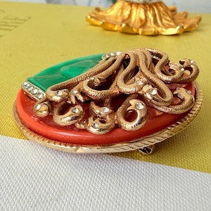 May include: A vintage brooch with a gold-tone metal frame. The brooch features a red and green oval base, with a gold-tone snake design and clear rhinestone accents. The brooch has a pin closure on the back.