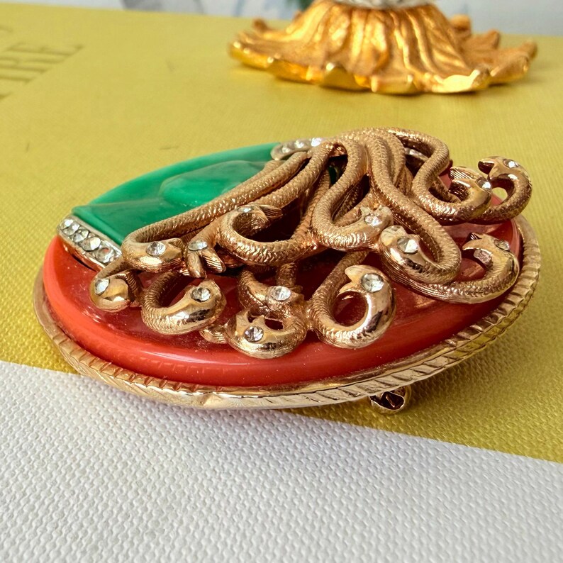 May include: A vintage brooch with a gold-tone metal frame. The brooch features a red and green oval base, with a gold-tone snake design and clear rhinestone accents. The brooch has a pin closure on the back.