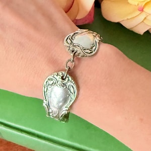 Puede incluir: Pulsera plateada con dos colgantes de cuchara ornamentados, unidos por una cadena. Las cucharas presentan diseños florales y de volutas detallados. La pulsera se lleva en la muñeca, mostrando la joyería de inspiración vintage.
