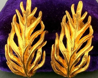 May include: A pair of gold-toned, leaf-shaped clip-on earrings. The earrings have a textured surface and a detailed, organic design. They are displayed on a purple velvet surface, showcasing their intricate craftsmanship and vintage style.